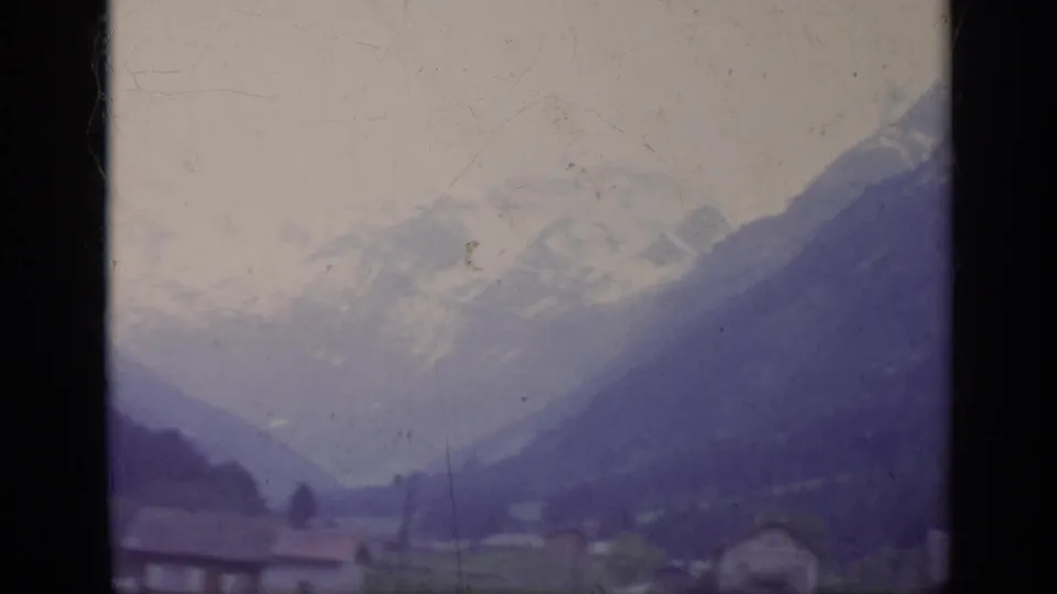 1962 Italy 8mm view down an alpine valley with small houses, utility poles, fields, and snow-covered mountains in the distance.