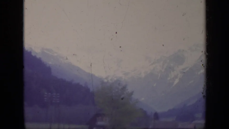 1962 Italy 8mm frame showing valley houses, dark wooded slopes, and snow-capped mountains softened by film grain and exposure.