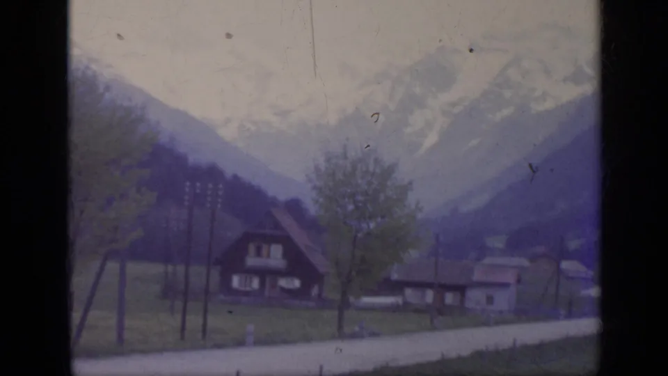 1962 8mm view from a passing train toward rural Italian village buildings below mountain slopes.
