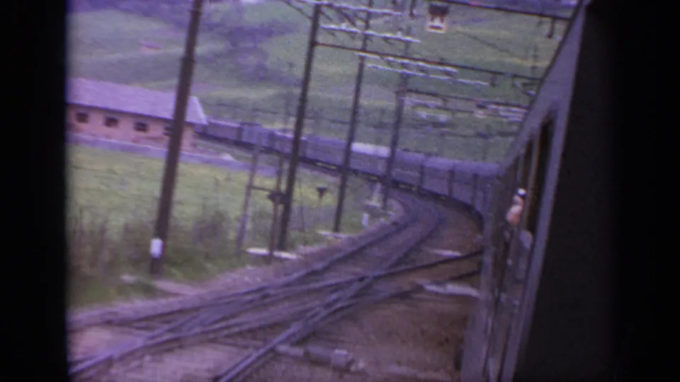 Train-side view of curving rails, power poles, and rural buildings in an Italian mountain valley.