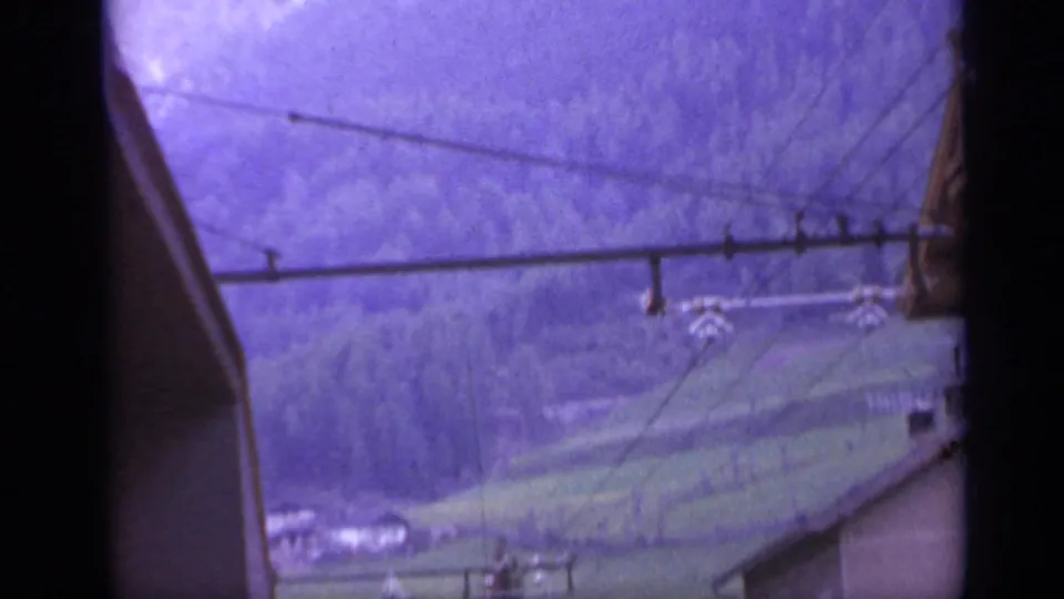 A train-window view catches overhead lines against wooded slopes and a green valley in rural Italy.