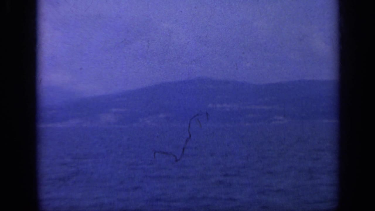 1962 Italy 8mm frame showing blue water, distant hazy mountains, and a shoreline under a pale sky.