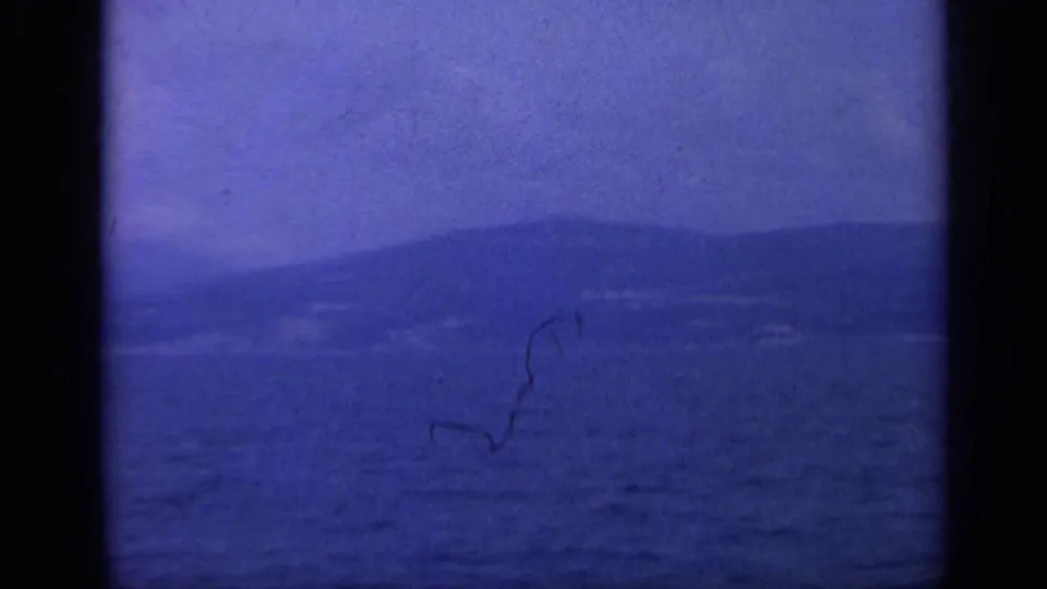 1962 Italy 8mm frame showing blue water, distant hazy mountains, and a shoreline under a pale sky.