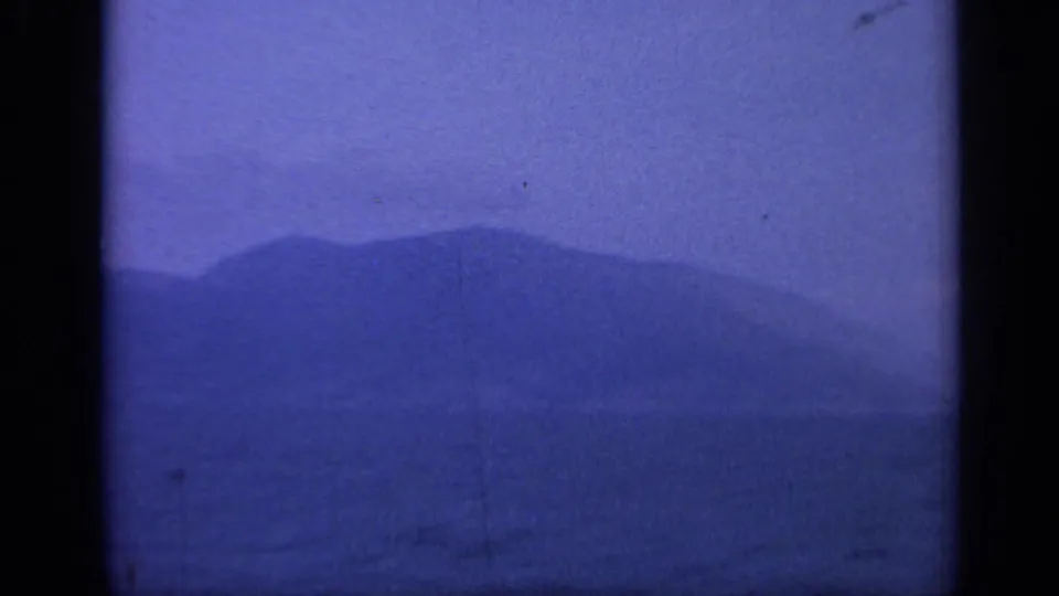 1962 Italy coastal view with a low mountain shape beyond blue water, softened by haze and film grain.