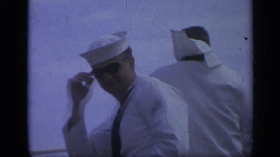 1962 Italy home-movie frame showing a sailor adjusting sunglasses beside another white-uniformed sailor aboard ship.