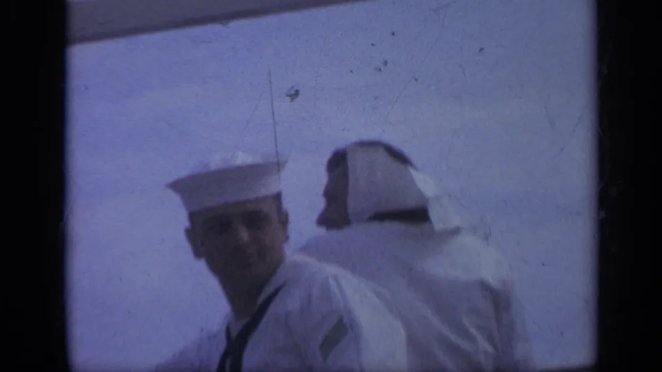 A sailor in a white cap looks toward the camera while another faces away during a 1962 shipboard scene near Italy.