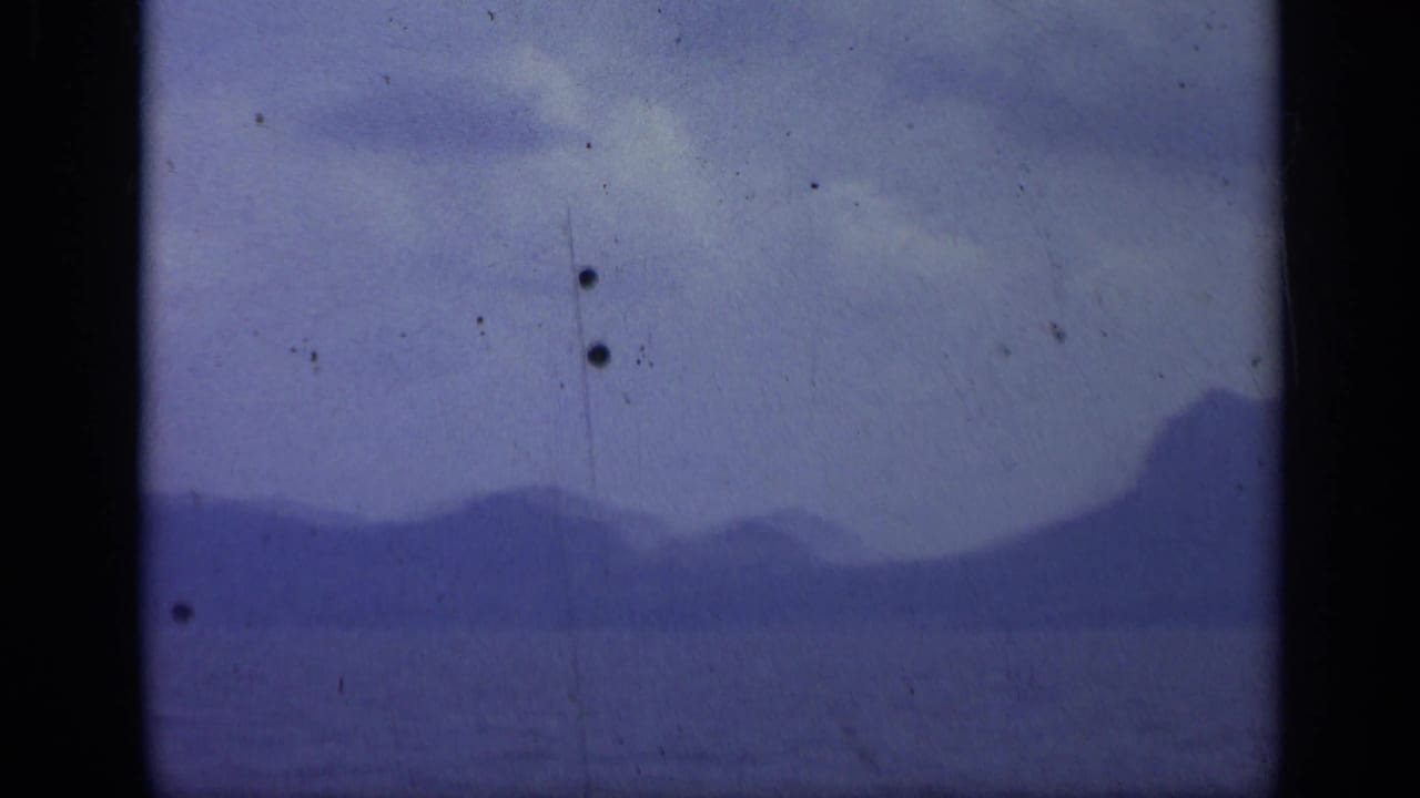 1962 Italian 8mm frame showing a distant island silhouette over calm sea, with cloudy sky and aged film marks.
