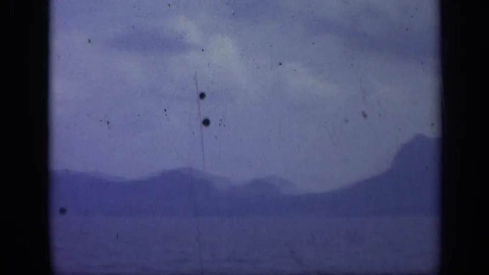 1962 Italian 8mm frame showing a distant island silhouette over calm sea, with cloudy sky and aged film marks.