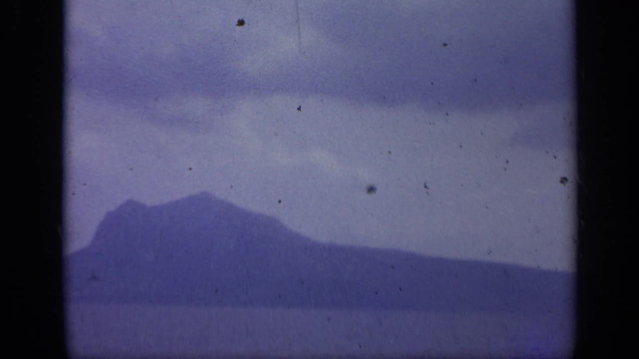 1962 Italian home-movie view of a mountainous island across calm water, softened by blue-purple film fading.