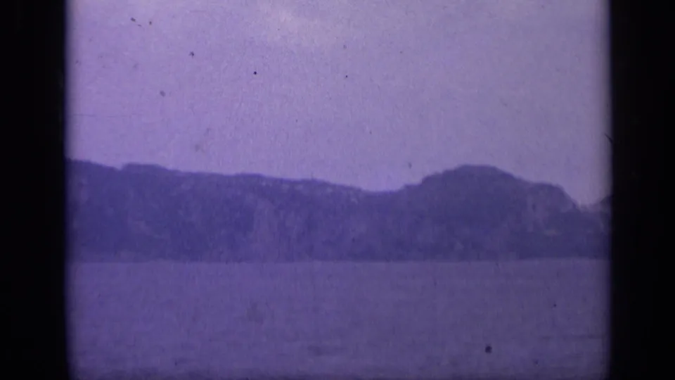 1962 Italian 8mm frame showing calm water and a low rocky shoreline beneath a hazy purple-tinted sky.