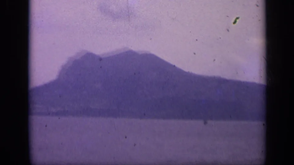 1962 Italian 8mm frame showing a large rocky landform across water, with visible dust and soft focus.