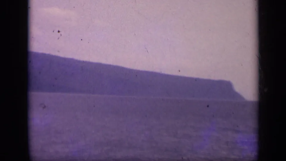 1962 Italian 8mm frame of a long rocky headland and calm water, tinted purple by the film scan.