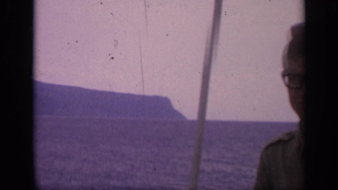 Sea and rocky headland seen through a dark film gate with a partially obscured adult face at center.