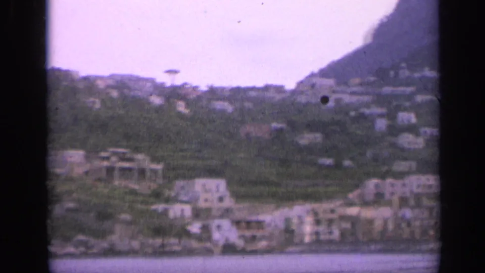 1962 Italy coastal town view with shoreline buildings, terraced hillside, mountain slope, and sea in the foreground.