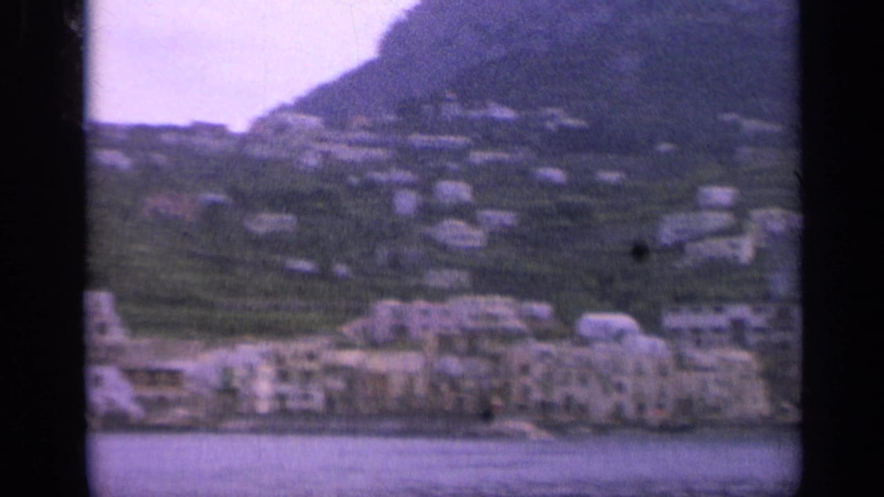 Distant Italian hillside village with pale buildings, terraced greenery, and mountains.