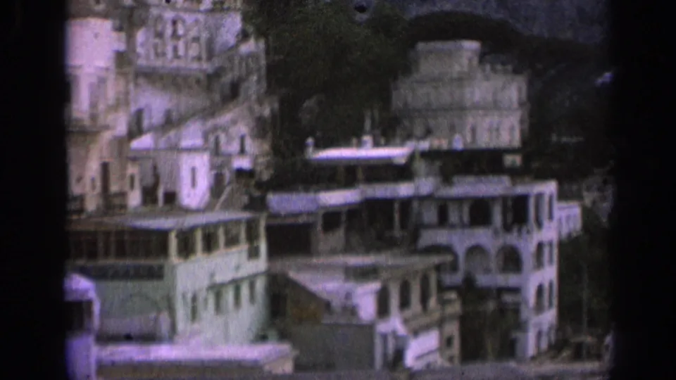 Closer 1962 8mm view of pale Italian hillside buildings with arches, balconies or roof edges, and rocky slopes behind.