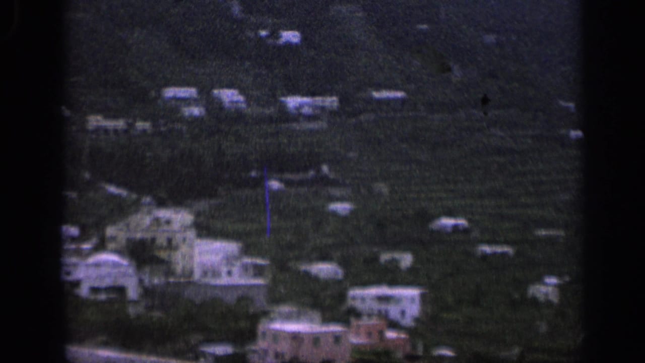 Cluster of white buildings on a green hillside seen from afar.