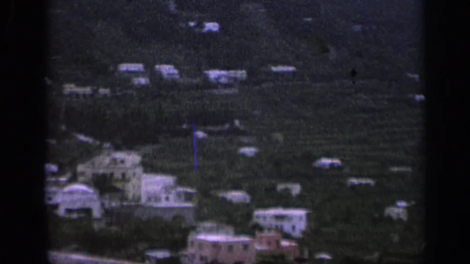 1962 Italy 8mm frame with a distant cluster of pale buildings across a dark green hillside.