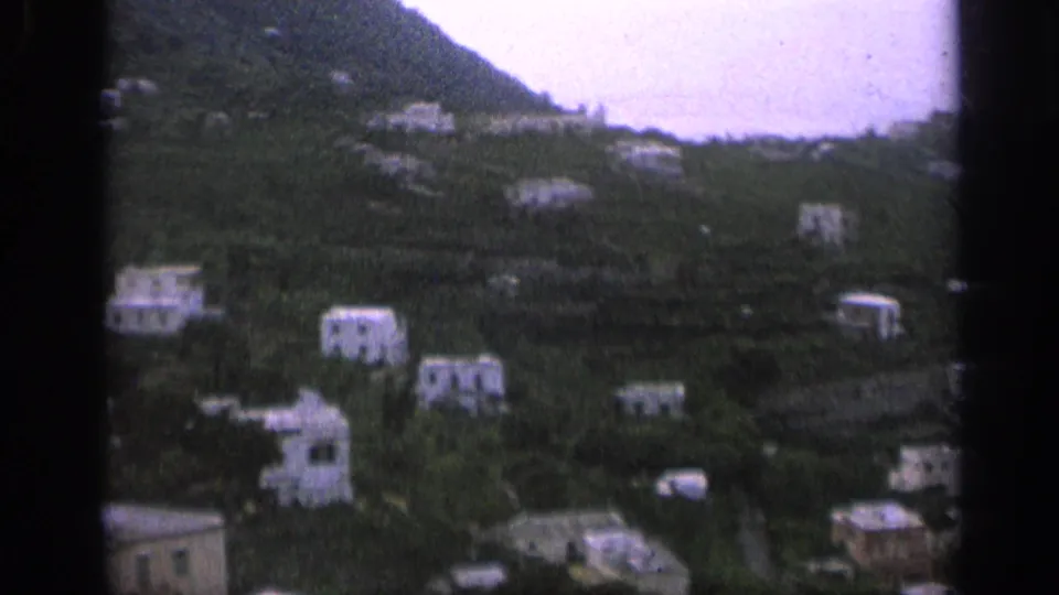1962 Italy 8mm frame of pale hillside houses below a ridge with a bright distant horizon.