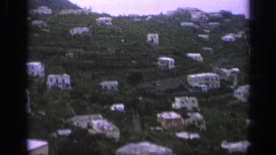 1962 Italy 8mm frame showing pale hillside houses set among dense greenery, viewed from a distance.