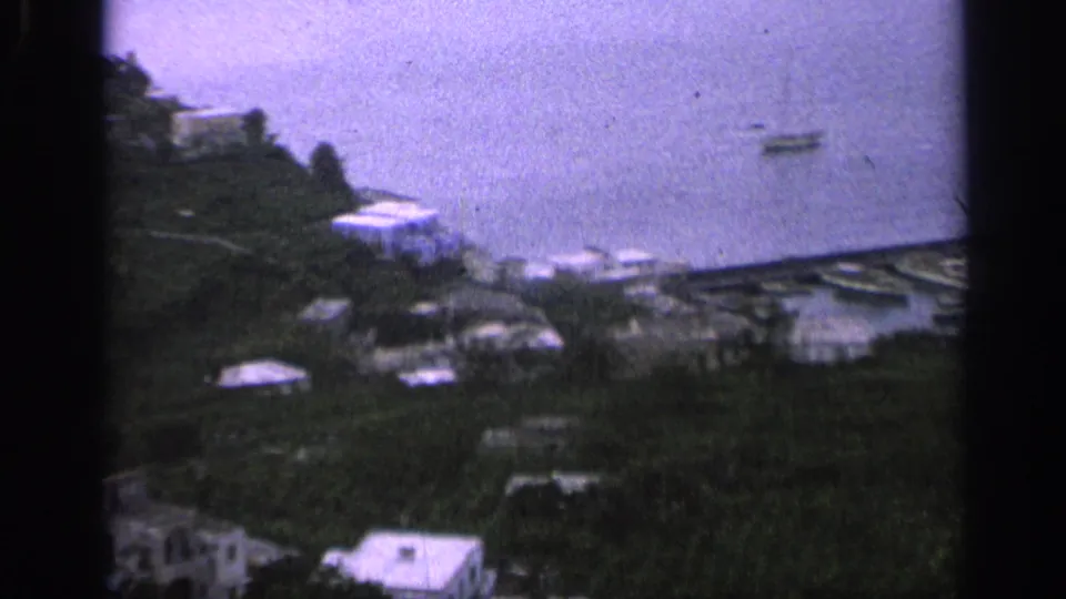 1962 Italy 8mm frame with hillside vegetation, scattered houses, shoreline, and a small harbor below.