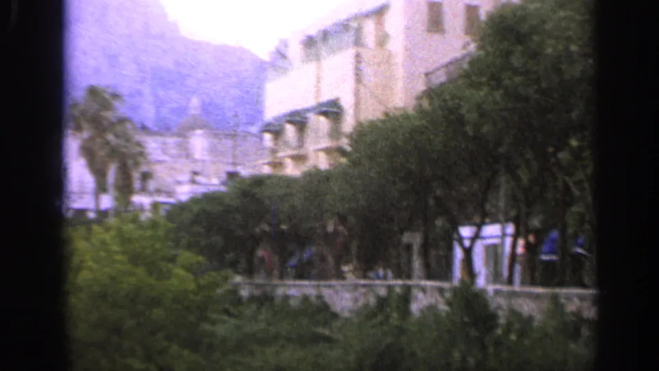 Italian town view with pale buildings, palm trees, garden greenery, and mountains in the background on 8mm film.