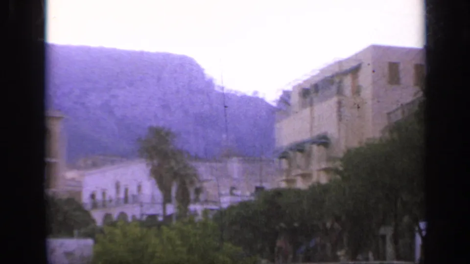 1962 Italy 8mm travel frame showing pale hillside buildings, palms, trees, and purple-hued mountains.