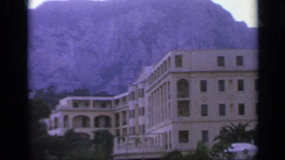 1962 Italy 8mm frame with pale buildings, arched balconies, trees, and a steep mountain behind them.