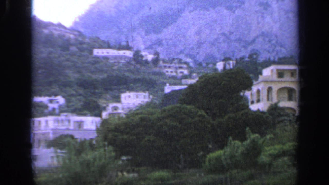 White hillside buildings among trees below a rocky cliff.