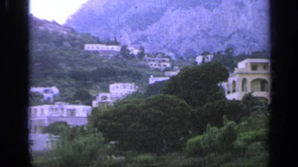 1962 Italy 8mm frame showing white buildings, dense greenery, and a rocky hillside seen through dark film-frame edges.