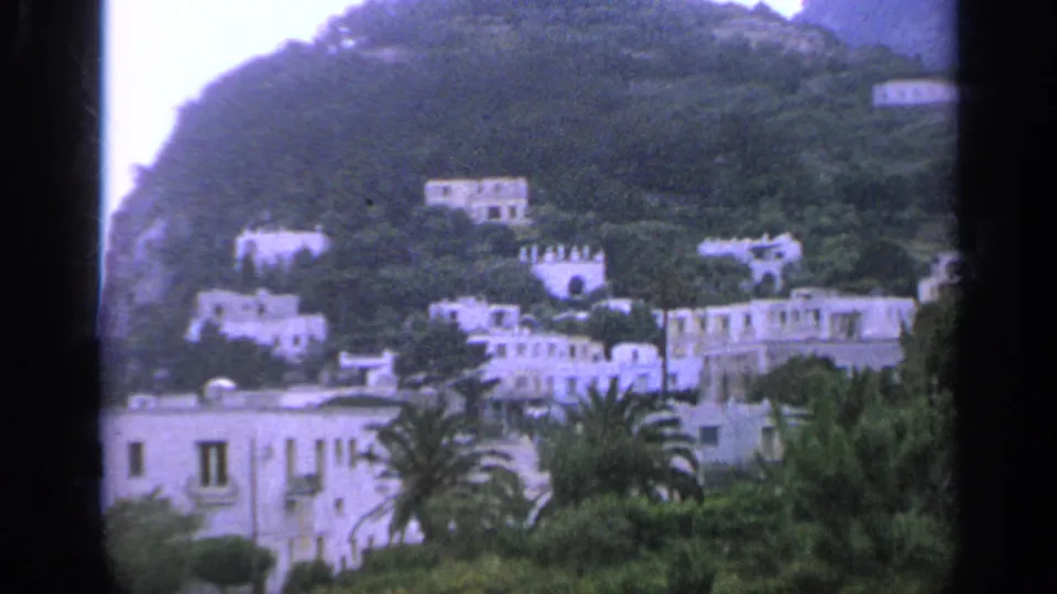 1962 Italy home-movie view of white hillside villas, palm trees, and thick foliage below a rocky slope.