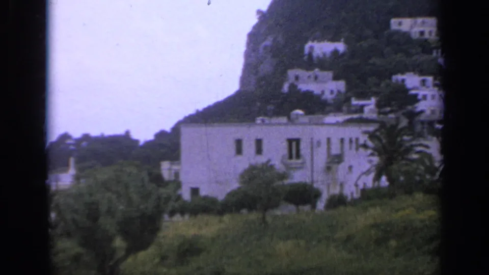 1962 Italy 8mm frame with a pale building, palm tree, shrubs, and white houses higher on the wooded hillside.