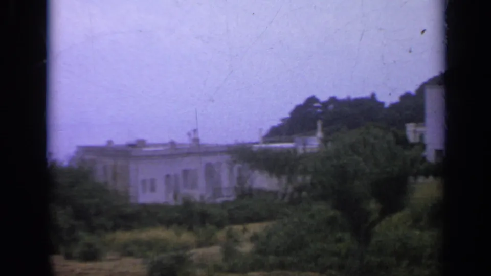 1962 Italy 8mm frame showing a low white building, trees, garden growth, and a hillside under a pale sky.