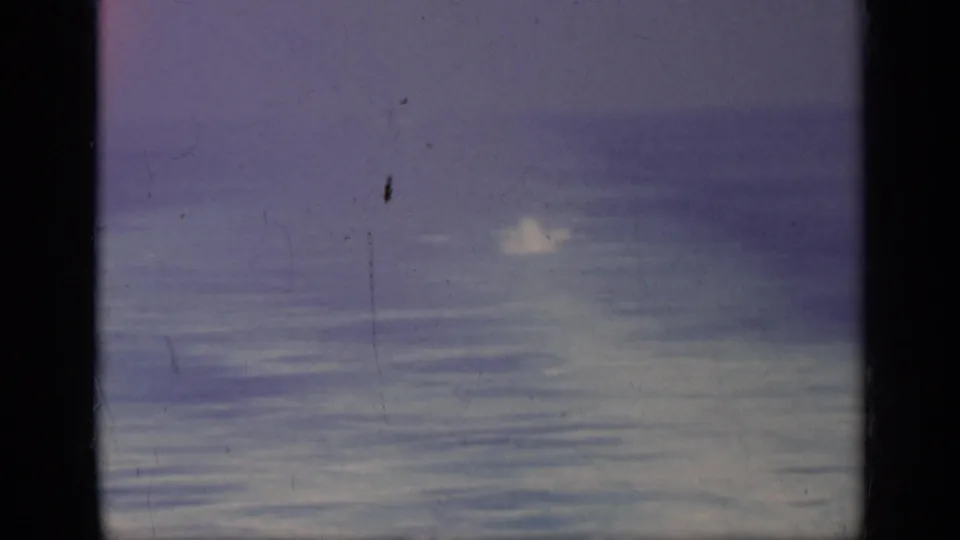 Purple-blue 8mm sea view from the 1962 Italy clip, showing a distant bright plume or splash on the water with dust, scratches, and frame edges.