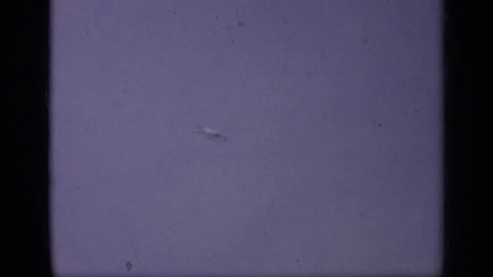 A distant airplane appears in a purple-tinted sky in 1962 Italy 8mm home-movie footage, with dust and soft focus visible.