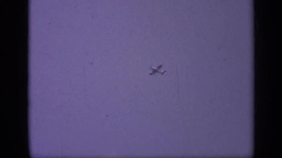 The airplane remains small and distant in the purple-tinted sky, surrounded by visible scratches, dust, and soft 8mm grain.