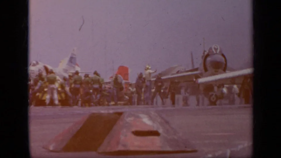 1962 Italy carrier-deck view with jets parked in line and sailors working around the aircraft under a pale sky.