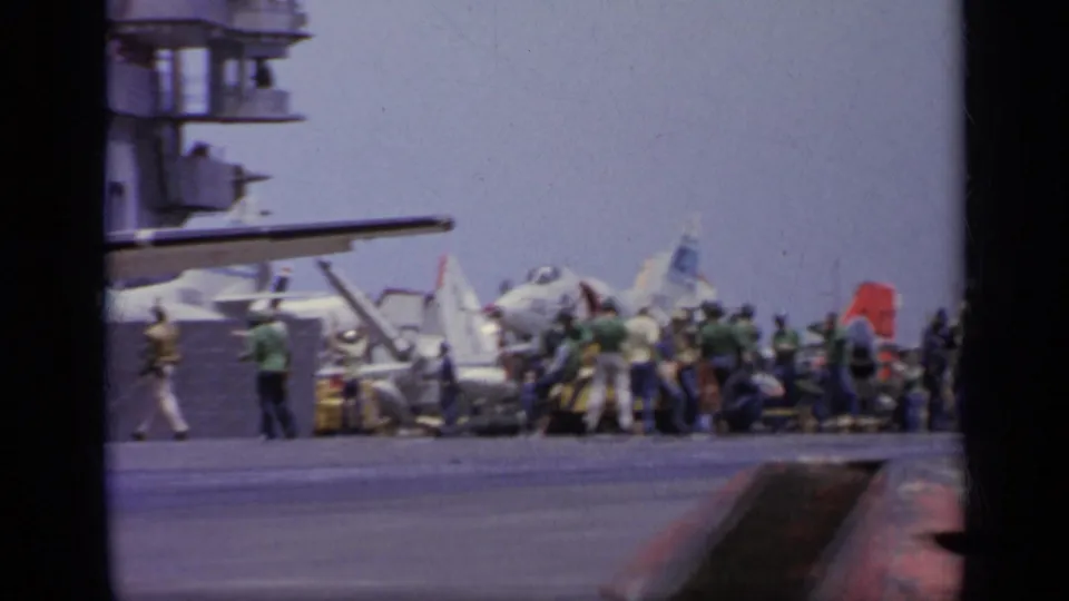 Wide 1962 8mm view of sailors in colored deck shirts working around military aircraft on a carrier deck near Italy.