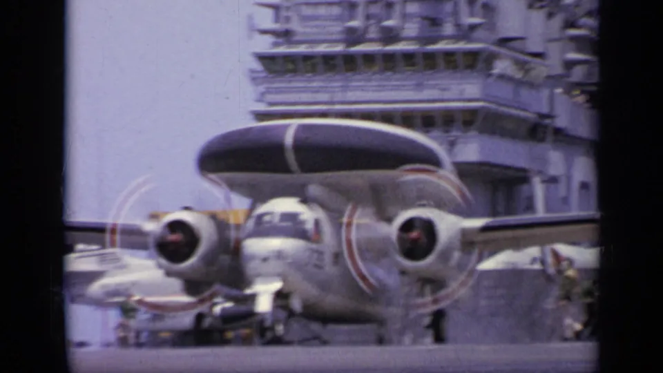 1962 carrier-deck frame showing a twin-engine radar aircraft in front of the carrier island, with sailors nearby.