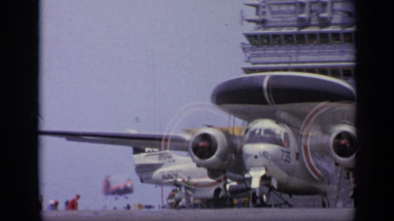 Radar-dome aircraft on an aircraft carrier deck with crew and island structure nearby.