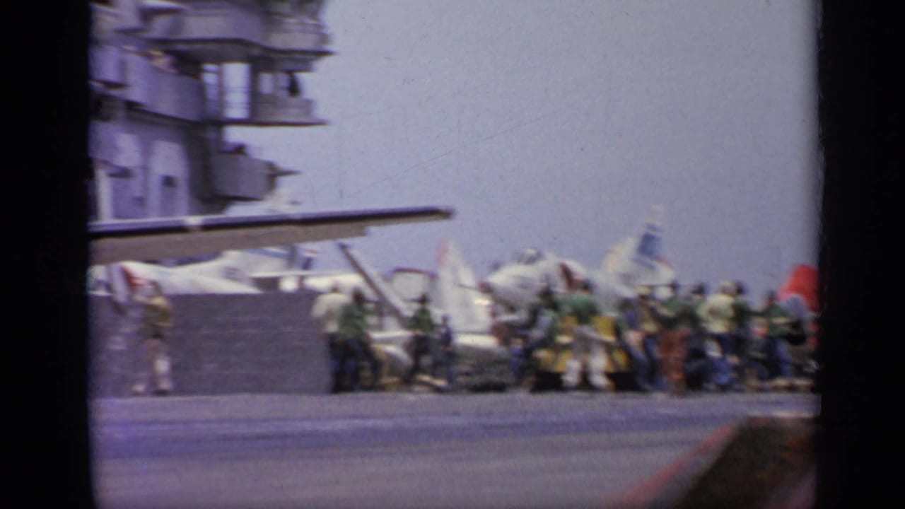 Carrier flight deck with parked jets, crew members, and island structure under clear sky.
