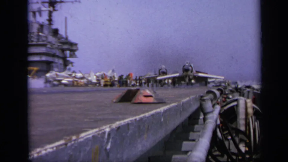 Wide 1962 8mm view along a U.S. Navy carrier deck near Italy, showing jets, crew clusters, deck edge, and surrounding water.