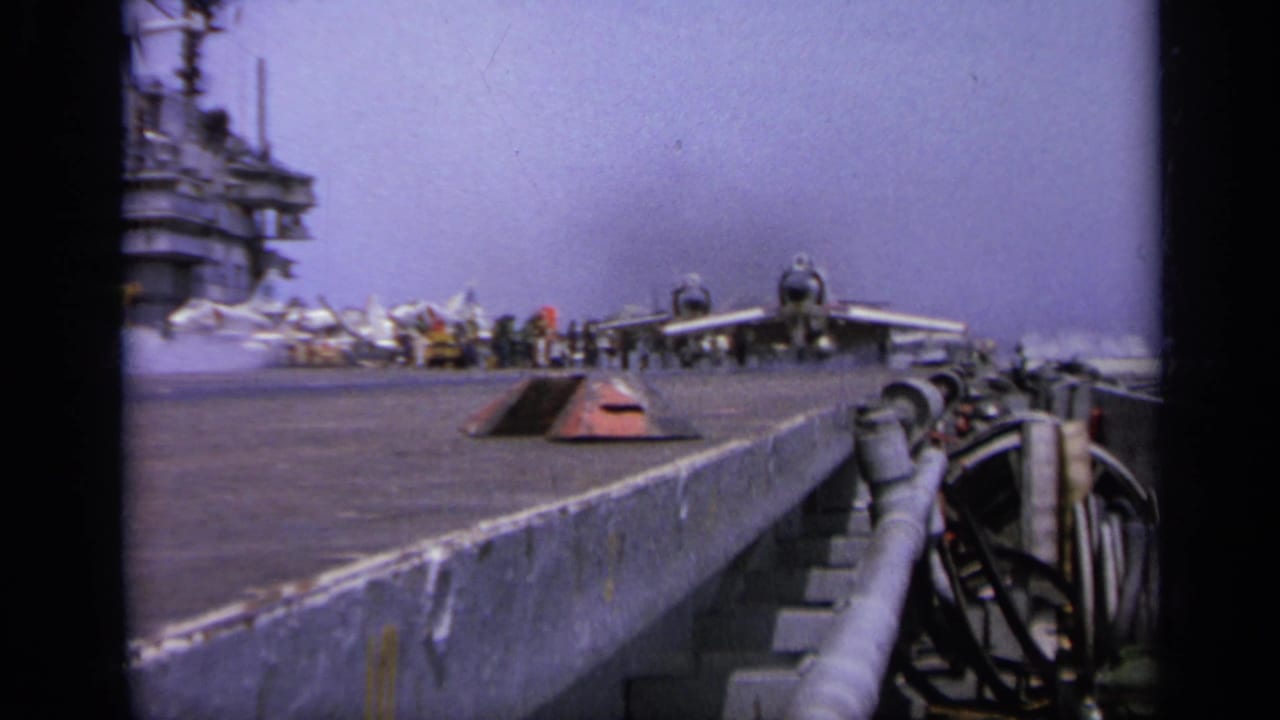Military jet on an aircraft carrier deck with crew and island structure in the background.