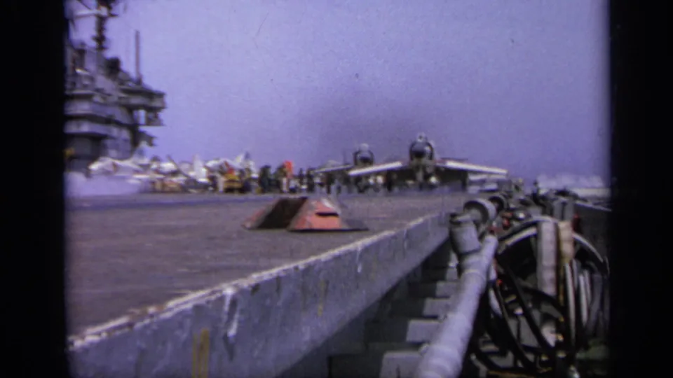 1962 Italy 8mm frame showing a military jet positioned on an aircraft carrier flight deck with deck crew and the island structure behind it.