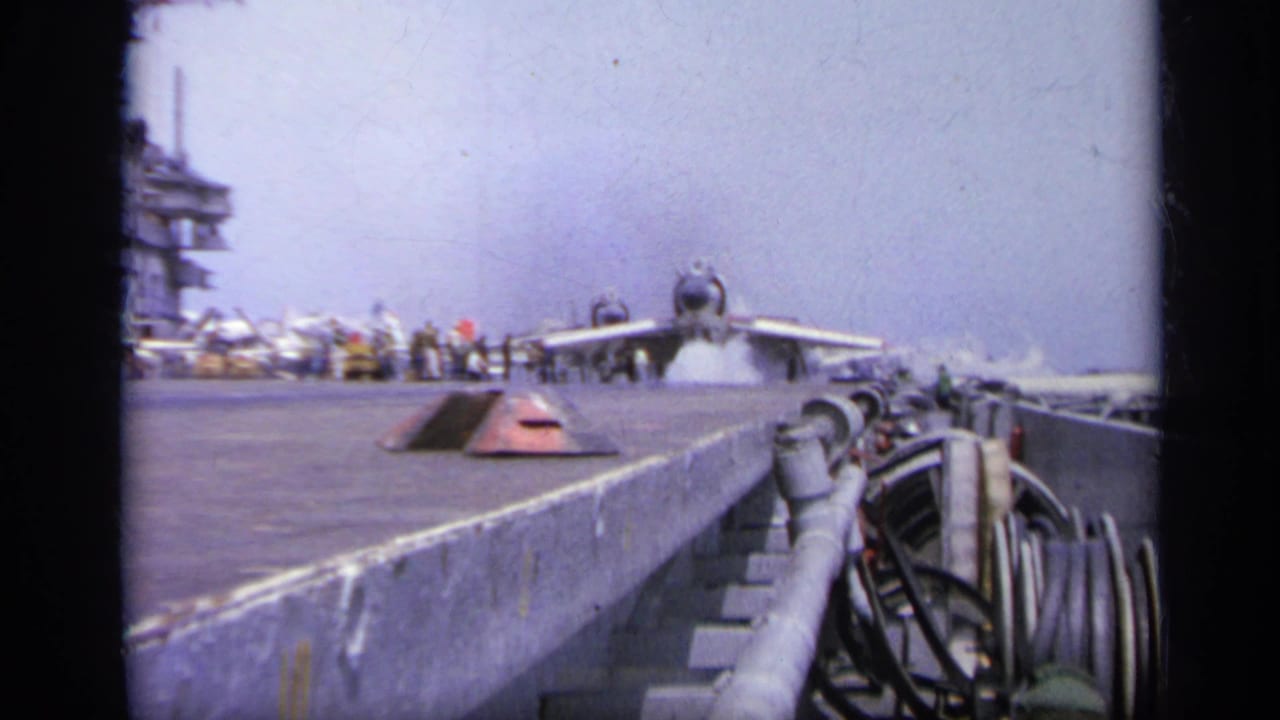 Jet aircraft seen from a low angle on a carrier flight deck beside deck-edge equipment.