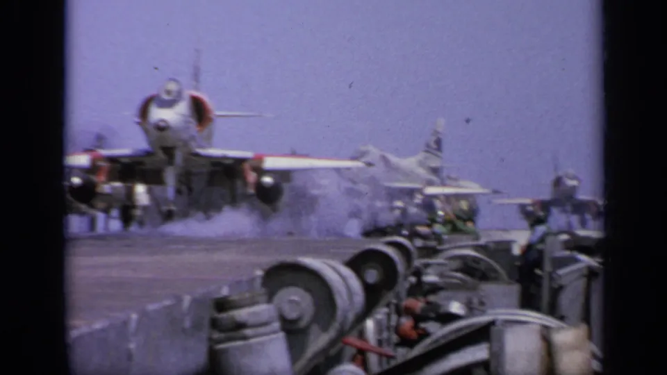 1962 Italy 8mm frame showing a military jet taking off or accelerating from an aircraft carrier deck, with deck machinery in the foreground.