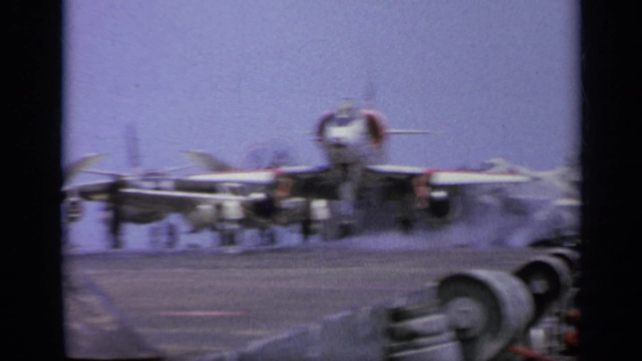 1962 Italy 8mm frame showing a jet on an aircraft carrier deck as exhaust haze gathers near sailors and equipment.