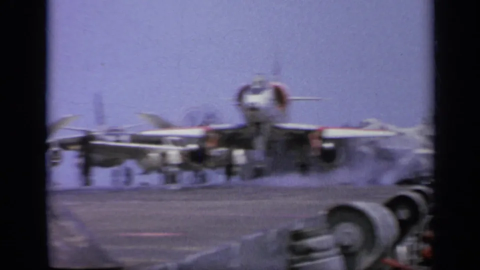 1962 Italy 8mm frame showing a jet on an aircraft carrier deck as exhaust haze gathers near sailors and equipment.