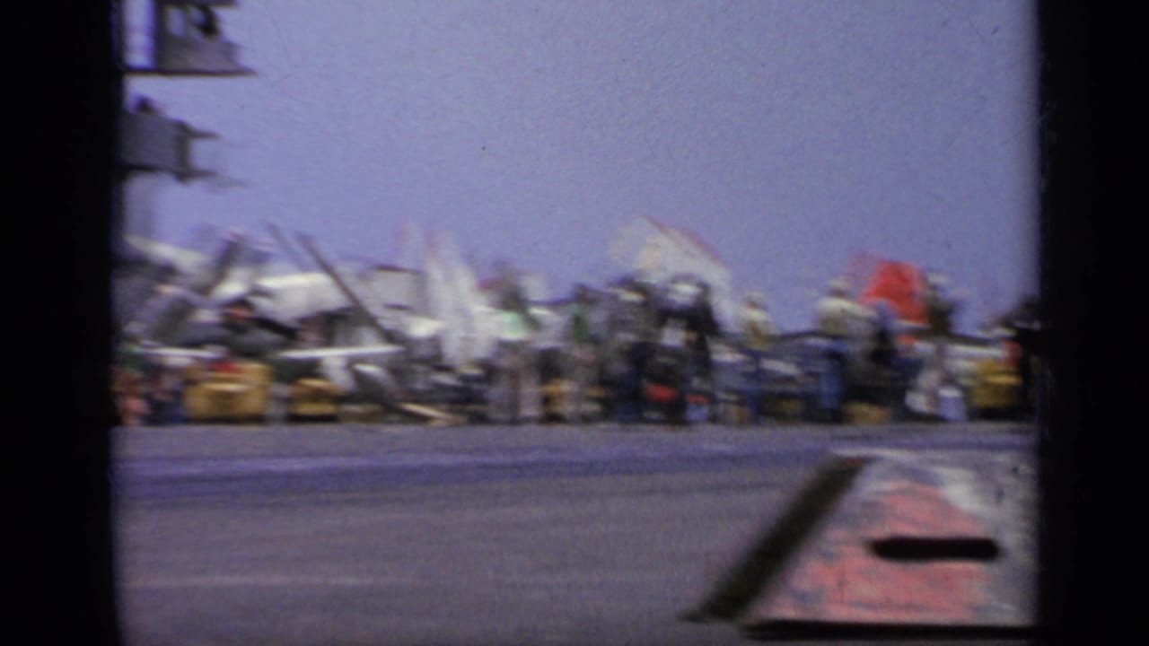 1962 Italy carrier-deck view with sailors gathered near parked aircraft, signal flags, and equipment under a bright sky.