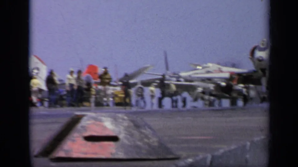 1962 Italy 8mm frame of sailors beside aircraft on a carrier deck, with signal flags and a deck structure in the foreground.